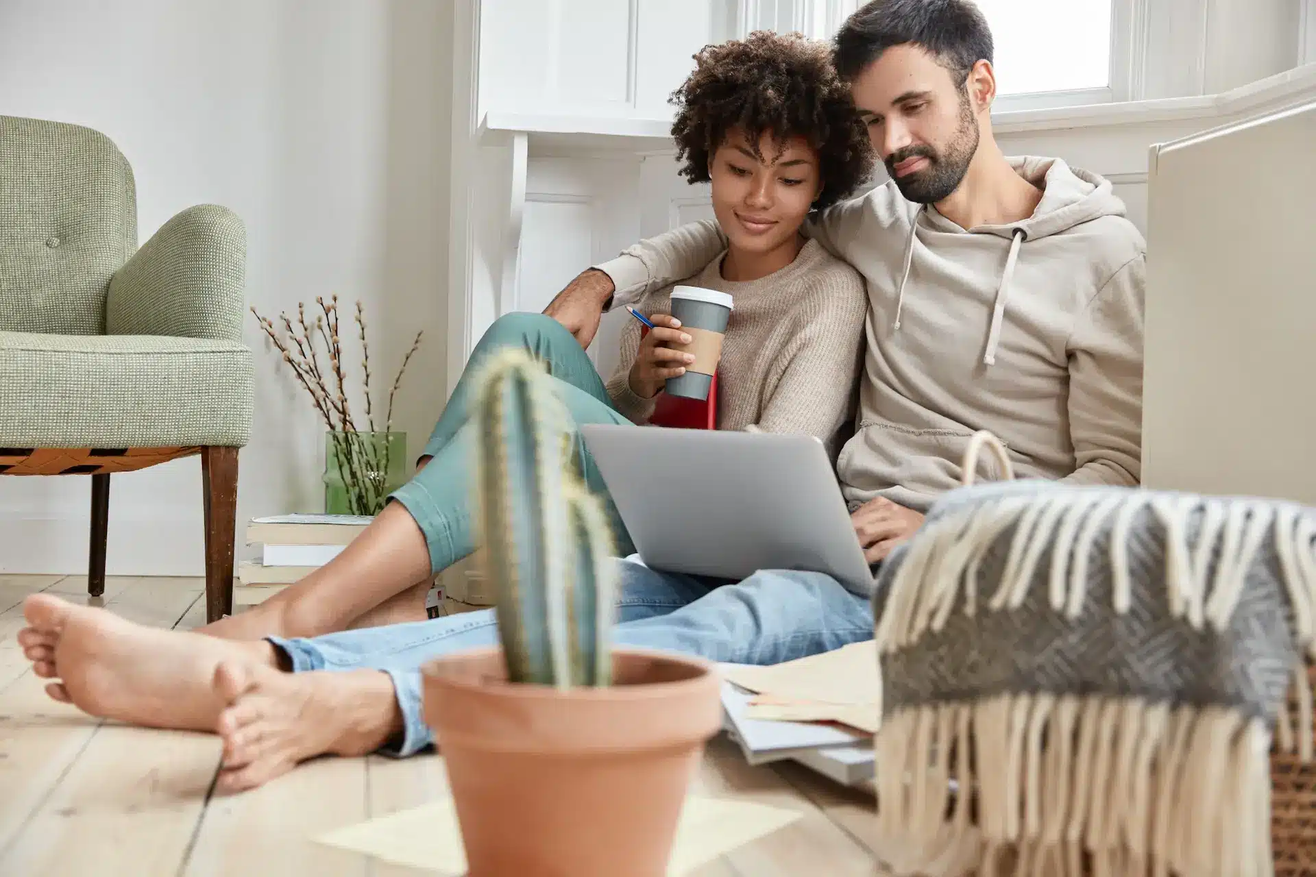 a couple sitting on the ground, looking at a laptop with a catcus in the foreground.